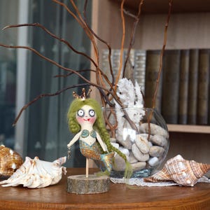 May include: A whimsical mermaid doll with green yarn hair, a gold crown, and a blue and gold tail stands on a wooden base. The doll is holding a small chain. Seashells and a glass vase with stones and branches are in the background.