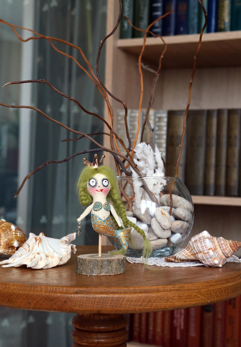 May include: A whimsical mermaid doll with green yarn hair, a gold crown, and a blue and gold tail stands on a wooden base. The doll is holding a small chain. Seashells and a glass vase with stones and branches are in the background.