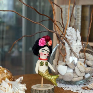 May include: A handmade mermaid doll with a black yarn hairstyle, floral headpiece, and a gold and green tail. The doll is mounted on a wooden base. A seashell and decorative vase are in the background.