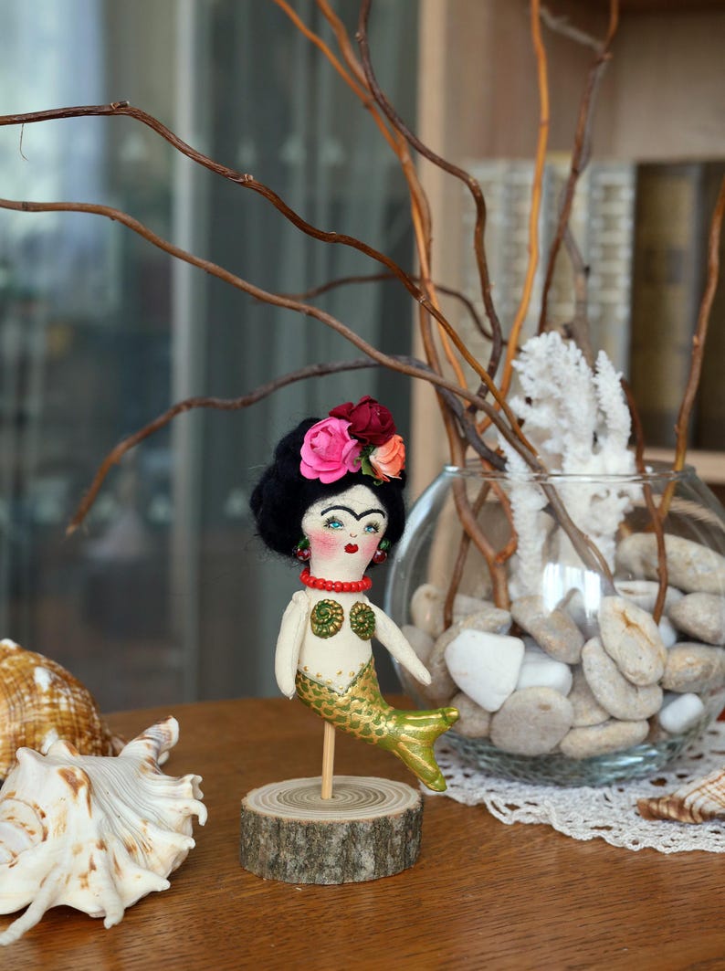 May include: A handmade mermaid doll with a black yarn hairstyle, floral headpiece, and a gold and green tail. The doll is mounted on a wooden base. A seashell and decorative vase are in the background.