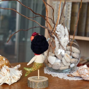 May include: A handmade mermaid doll with a black yarn hair, a red necklace, and a green and gold tail. The doll is mounted on a wooden base. Seashells and a glass bowl with stones and branches are in the background.