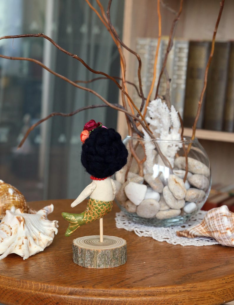 May include: A handmade mermaid doll with a black yarn hair, a red necklace, and a green and gold tail. The doll is mounted on a wooden base. Seashells and a glass bowl with stones and branches are in the background.
