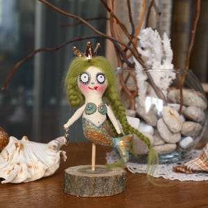 May include: A whimsical mermaid doll with green yarn hair, a gold crown, and a blue and gold tail. The doll is mounted on a wooden base and holds a small charm. The doll is a unique handmade item.