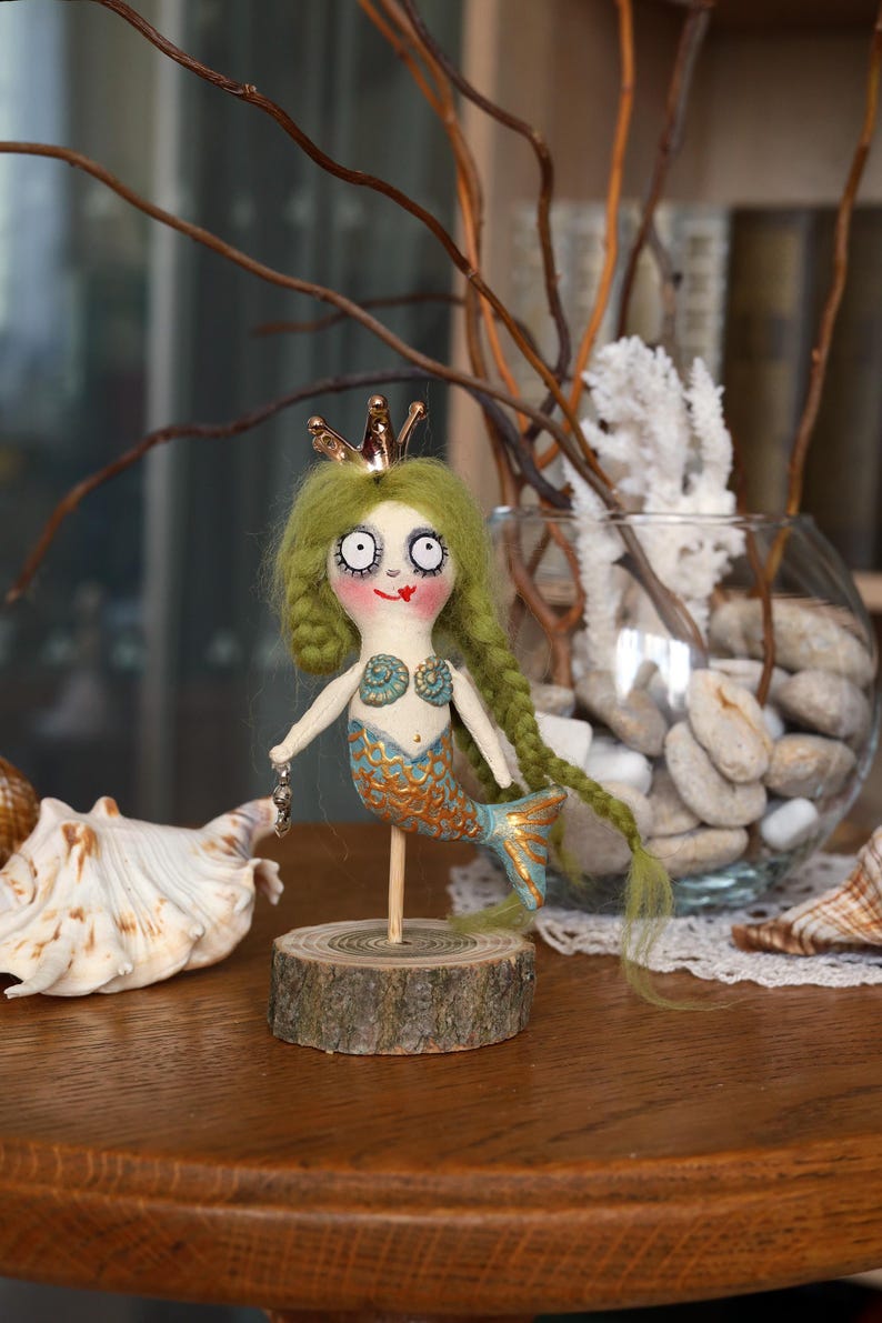 May include: A whimsical mermaid doll with green yarn hair, a gold crown, and a blue and gold tail. The doll is mounted on a wooden base and holds a small charm. The doll is a unique handmade item.