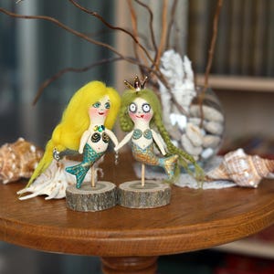 May include: Two handmade mermaid dolls with yellow and green hair, holding hands, stand on wooden bases. They have teal and gold tails, and are decorated with small gems. The dolls are displayed on a wooden table with seashells and branches.