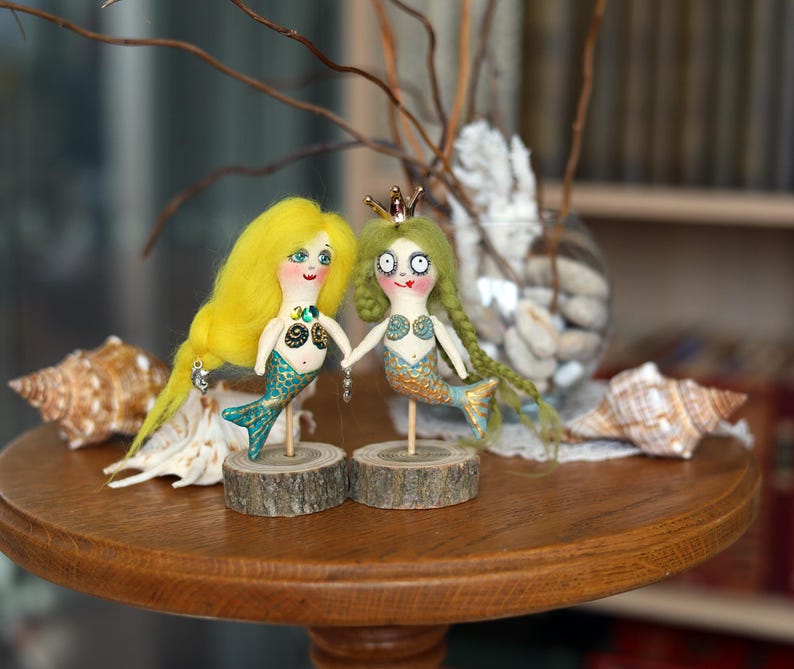May include: Two handmade mermaid dolls with yellow and green hair, holding hands, stand on wooden bases. They have teal and gold tails, and are decorated with small gems. The dolls are displayed on a wooden table with seashells and branches.