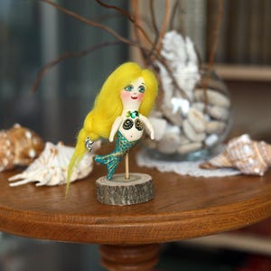 May include: A handmade mermaid doll with yellow hair, a turquoise tail, and a wooden base. The doll is on a round wooden table with seashells and a glass vase in the background. The mermaid doll is approximately 5 inches tall.