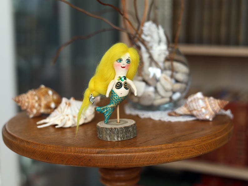 May include: A handmade mermaid doll with yellow hair, a turquoise tail, and a wooden base. The doll is on a round wooden table with seashells and a glass vase in the background. The mermaid doll is approximately 5 inches tall.