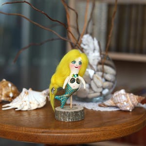 May include: A handmade mermaid doll with yellow hair, blue eyes, and a teal tail. The doll is mounted on a small wooden base. Seashells and decorative branches are in the background, creating a whimsical scene.