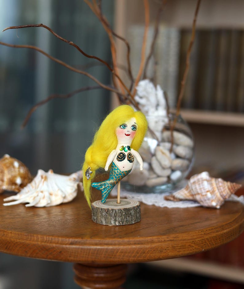 May include: A handmade mermaid doll with yellow hair, blue eyes, and a teal tail. The doll is mounted on a small wooden base. Seashells and decorative branches are in the background, creating a whimsical scene.