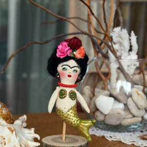 May include: A handmade mermaid doll with a black hairstyle, floral headpiece, and a gold and green tail. The doll is mounted on a wooden base. The doll has a red beaded necklace and earrings. The doll is a unique decorative item.