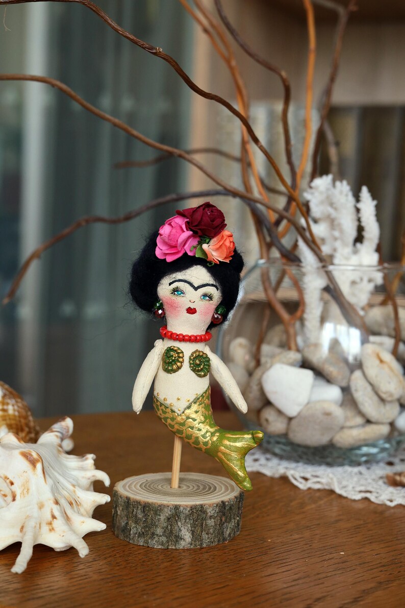 May include: A handmade mermaid doll with a black hairstyle, floral headpiece, and a gold and green tail. The doll is mounted on a wooden base. The doll has a red beaded necklace and earrings. The doll is a unique decorative item.