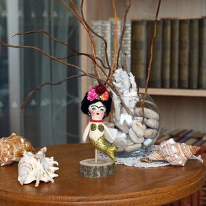 May include: A handmade mermaid doll with a black wig and floral crown stands on a wooden base. The doll has a gold tail and a painted face. Seashells and a glass vase with branches and stones are on the table.