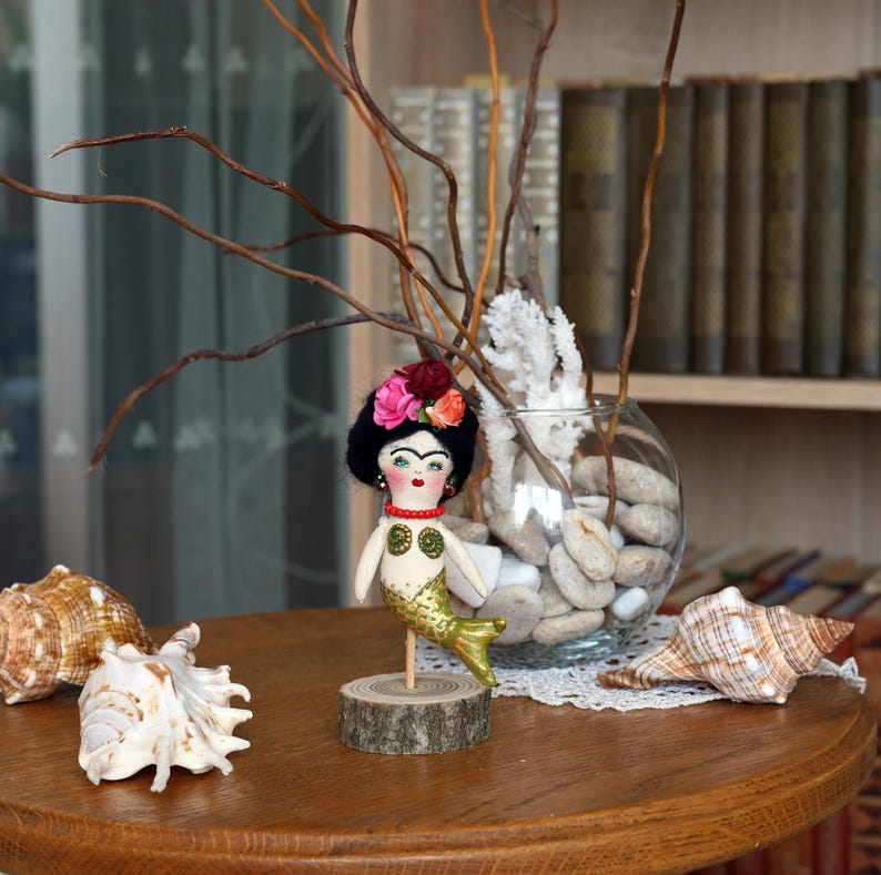 May include: A handmade mermaid doll with a black wig and floral crown stands on a wooden base. The doll has a gold tail and a painted face. Seashells and a glass vase with branches and stones are on the table.