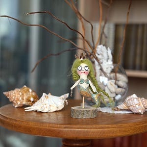 May include: A whimsical mermaid doll with green hair and a gold crown stands on a wooden base. The doll has a blue and gold tail and a seashell bra. Seashells and a glass vase with decorative elements are on a wooden table.