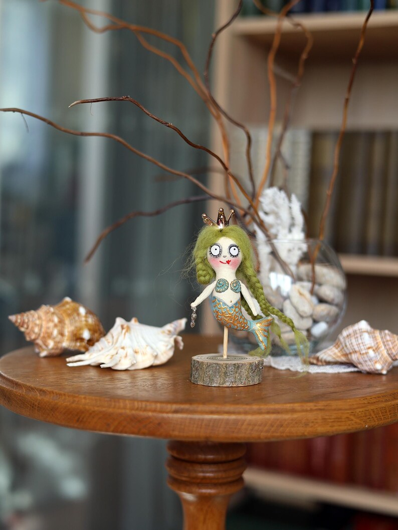 May include: A whimsical mermaid doll with green hair and a gold crown stands on a wooden base. The doll has a blue and gold tail and a seashell bra. Seashells and a glass vase with decorative elements are on a wooden table.
