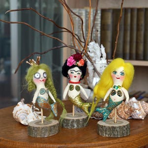 May include: Three handmade mermaid dolls with unique features. One doll has green hair and a gold crown, another has black hair with a floral headpiece, and the third has yellow hair. Each doll has a mermaid tail and is mounted on a wooden base.