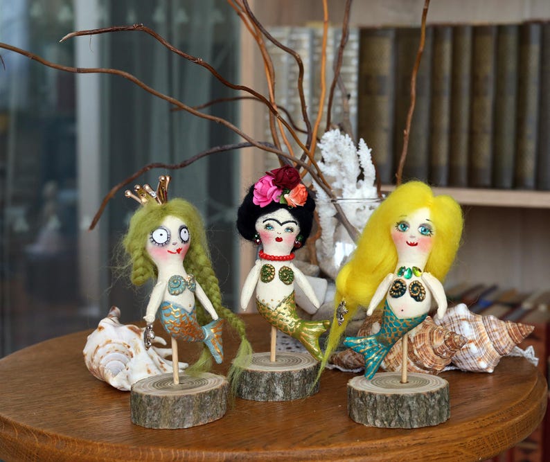 May include: Three handmade mermaid dolls with unique features. One doll has green hair and a gold crown, another has black hair with a floral headpiece, and the third has yellow hair. Each doll has a mermaid tail and is mounted on a wooden base.