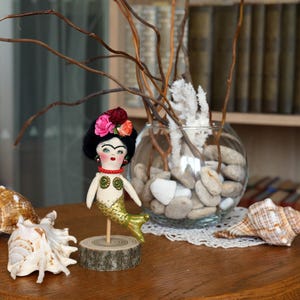 May include: A handmade mermaid doll with a black wig, floral headpiece, and gold tail. The doll is mounted on a wooden base. Seashells and a glass bowl with stones and branches are in the background.