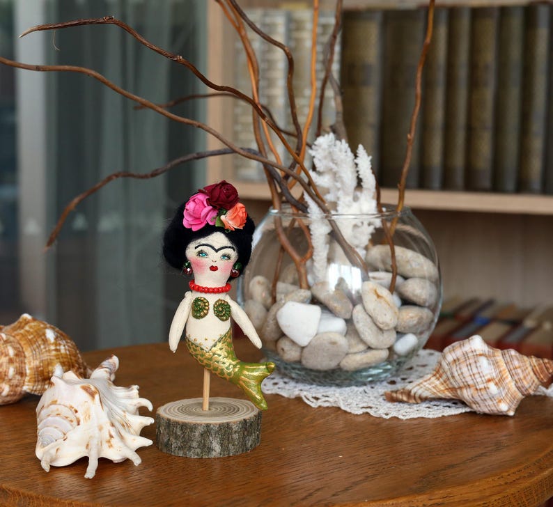 May include: A handmade mermaid doll with a black wig, floral headpiece, and gold tail. The doll is mounted on a wooden base. Seashells and a glass bowl with stones and branches are in the background.