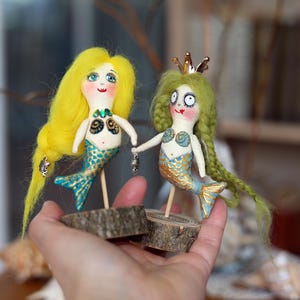 May include: Two handmade mermaid dolls. One has yellow hair and a turquoise tail, the other has green hair and a gold crown. Both dolls are holding hands and mounted on wooden bases. The dolls have painted faces and are holding small silver charms.