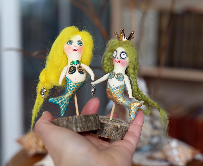 May include: Two handmade mermaid dolls. One has yellow hair and a turquoise tail, the other has green hair and a gold crown. Both dolls are holding hands and mounted on wooden bases. The dolls have painted faces and are holding small silver charms.