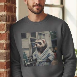 May include: Dark grey sweatshirt featuring a humorous image of a ferret in a bathrobe smoking a cigarette. The ferret is in front of a trailer. The sweatshirt is a solid dark grey color.