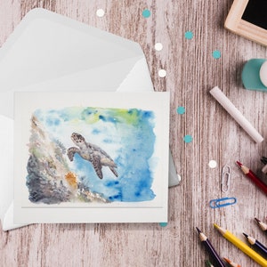 May include: A watercolor greeting card featuring a sea turtle swimming near a coral reef. The card is inside a white envelope, with pencils, a notepad, and a chalkboard on a wooden surface. The artwork has blue, green, and brown tones.