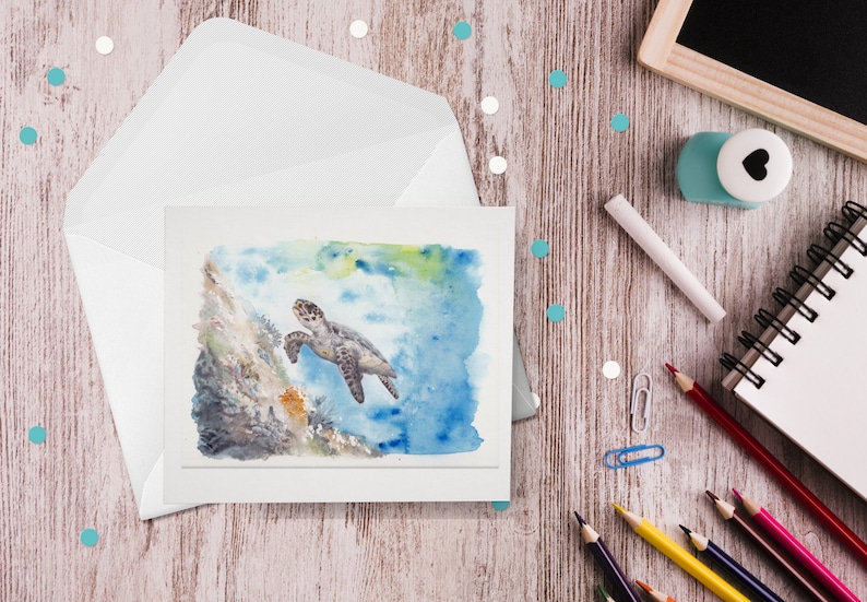 May include: A watercolor greeting card featuring a sea turtle swimming near a coral reef. The card is inside a white envelope, with pencils, a notepad, and a chalkboard on a wooden surface. The artwork has blue, green, and brown tones.