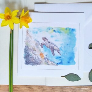 May include: A watercolor painting of a sea turtle swimming in blue water, presented on a white card. The card is on a notebook with a blue pen and yellow daffodils. Green leaves are scattered around the card.