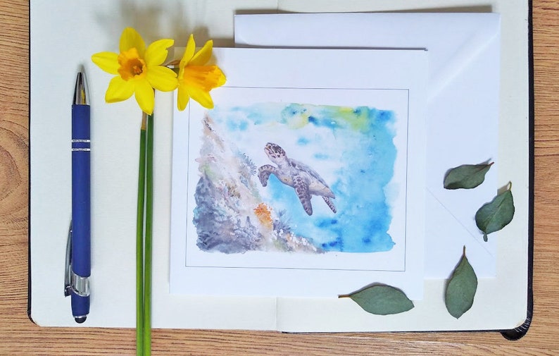 May include: A watercolor painting of a sea turtle swimming in blue water, presented on a white card. The card is on a notebook with a blue pen and yellow daffodils. Green leaves are scattered around the card.