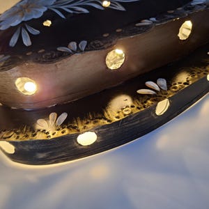Puede incluir: Una pieza decorativa de madera ovalada de dos niveles e iluminada. La superficie marr&oacute;n oscuro est&aacute; adornada con dise&ntilde;os florales blancos y azul claro y agujeros recortados que emiten un brillo c&aacute;lido. Los bordes est&aacute;n adornados con oro.