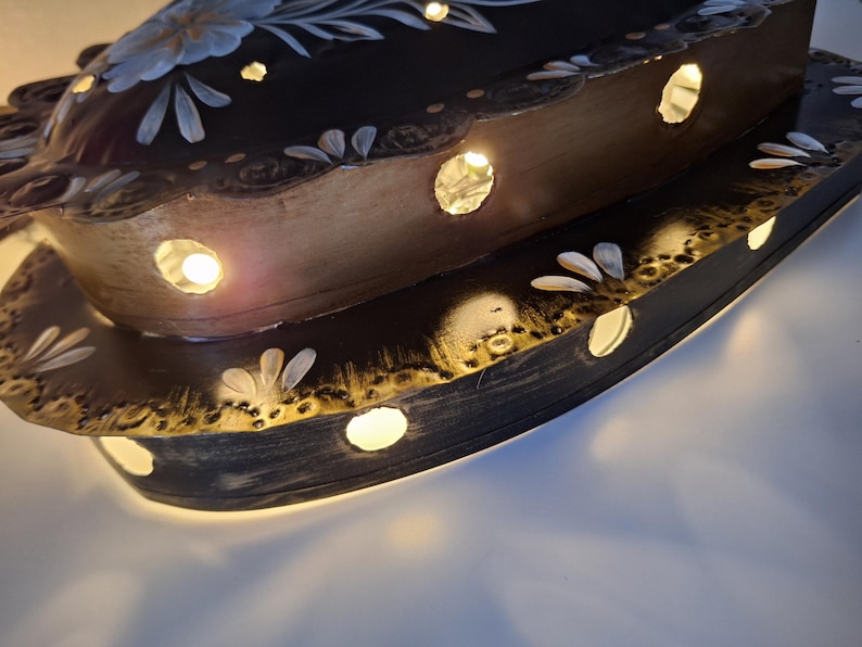 Puede incluir: Una pieza decorativa de madera ovalada de dos niveles e iluminada. La superficie marr&oacute;n oscuro est&aacute; adornada con dise&ntilde;os florales blancos y azul claro y agujeros recortados que emiten un brillo c&aacute;lido. Los bordes est&aacute;n adornados con oro.