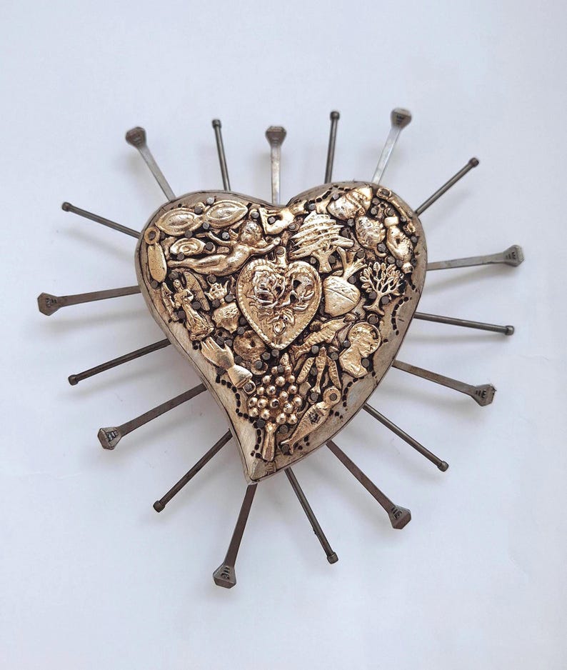 Puede incluir: Un adorno de pared en forma de coraz&oacute;n con acabado en oro met&aacute;lico, que presenta figuras en relieve que incluyen un coraz&oacute;n m&aacute;s peque&ntilde;o, un &aacute;rbol, uvas y un &aacute;ngel. El coraz&oacute;n est&aacute; rodeado por numerosas p&uacute;as met&aacute;licas oscuras que irradian hacia afuera.