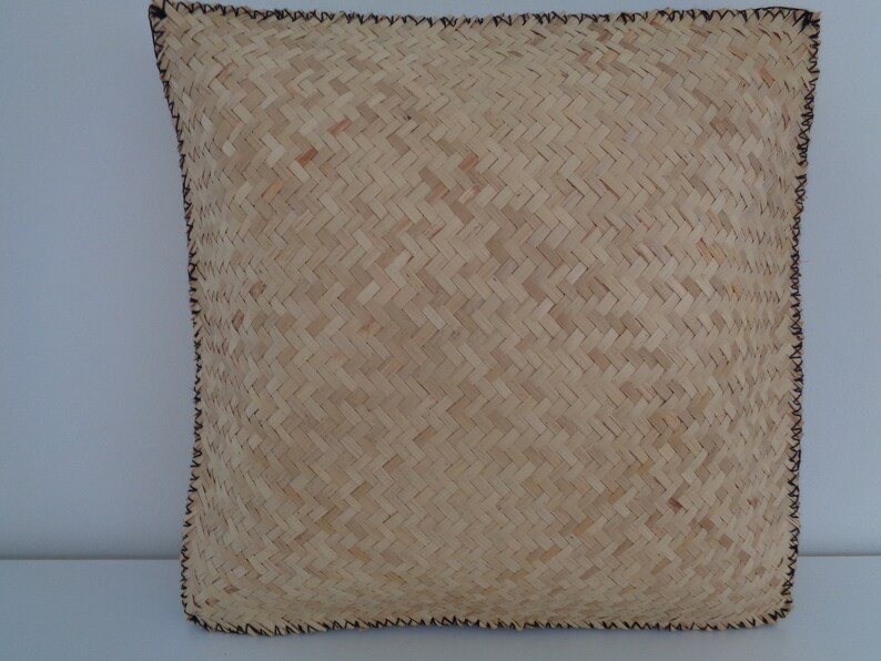 May include: A natural woven cushion with a brown stitched border. The cushion is made of a light brown material with a woven pattern.