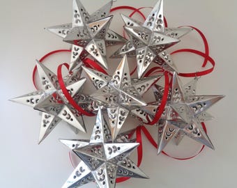 May include: A collection of six silver-colored star-shaped ornaments with intricate cut-out patterns on each point. Each star features a reflective metallic finish and is attached to a thin, bright red ribbon for hanging purposes.