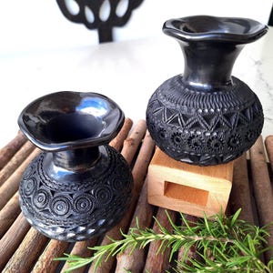 May include: Two black ceramic vases with intricate carved patterns and flared rims. One vase rests on a wooden block, while both sit on a bed of small wooden sticks. A sprig of green rosemary adds a natural touch.