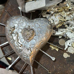 Puede incluir: Un adorno met&aacute;lico en forma de coraz&oacute;n de tono plateado con patrones circulares en relieve y un dije central. El adorno est&aacute; atravesado por varias varillas met&aacute;licas delgadas que irradian hacia afuera, con una peque&ntilde;a figura en el lado izquierdo.
