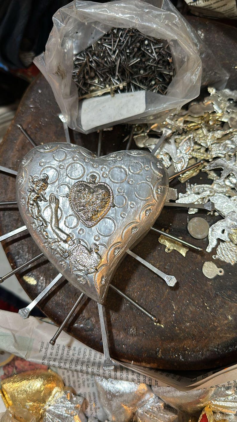 Puede incluir: Un adorno met&aacute;lico en forma de coraz&oacute;n de tono plateado con patrones circulares en relieve y un dije central. El adorno est&aacute; atravesado por varias varillas met&aacute;licas delgadas que irradian hacia afuera, con una peque&ntilde;a figura en el lado izquierdo.