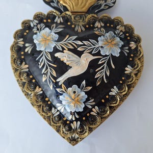 Puede incluir: Una pieza decorativa en forma de coraz&oacute;n con fondo negro, adornada con un colibr&iacute; y un dise&ntilde;o floral. Los bordes est&aacute;n detallados con detalles dorados. El colibr&iacute; es blanco y las flores son blancas y celeste.