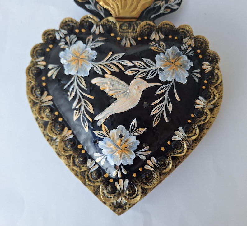 Puede incluir: Una pieza decorativa en forma de coraz&oacute;n con fondo negro, adornada con un colibr&iacute; y un dise&ntilde;o floral. Los bordes est&aacute;n detallados con detalles dorados. El colibr&iacute; es blanco y las flores son blancas y celeste.