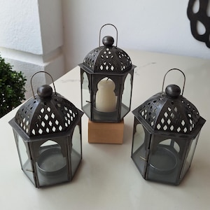 May include: Three black metal candle lanterns with intricate cut-out designs and glass panels. The lanterns are on a white surface.