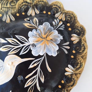 Puede incluir: Art&iacute;culo decorativo negro en forma de coraz&oacute;n con dise&ntilde;os florales y de colibr&iacute;es dorados y blancos. Los bordes est&aacute;n festoneados y pintados con detalles dorados y blancos. El art&iacute;culo tiene un acabado brillante.