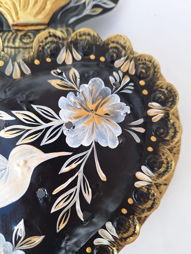 Puede incluir: Art&iacute;culo decorativo negro en forma de coraz&oacute;n con dise&ntilde;os florales y de colibr&iacute;es dorados y blancos. Los bordes est&aacute;n festoneados y pintados con detalles dorados y blancos. El art&iacute;culo tiene un acabado brillante.