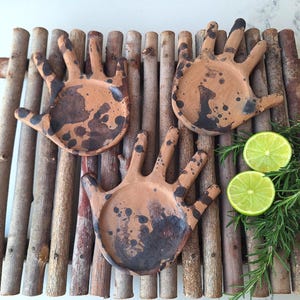 Puede incluir: Tres platos de terracota con forma de mano y salpicaduras de pintura negra descansan sobre una bandeja de madera. Dos mitades de lima y ramitas de romero están dispuestas a la derecha, añadiendo un elemento natural.