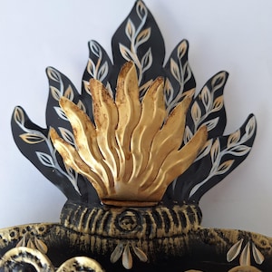 Puede incluir: Una pieza decorativa de metal con un dise&ntilde;o de llama estilizada. Las llamas son doradas, con detalles en negro y dorado. La pieza tiene una base detallada y ornamentada con elementos florales, y probablemente es un adorno de pared.