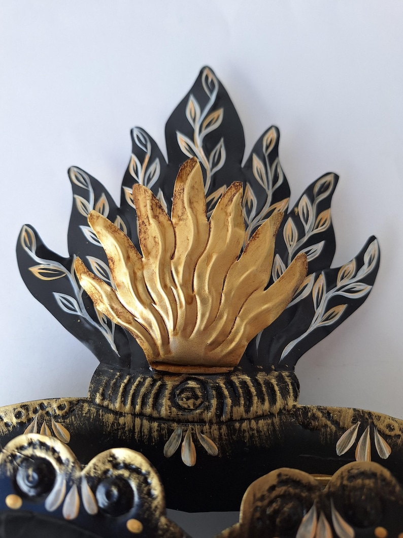 Puede incluir: Una pieza decorativa de metal con un dise&ntilde;o de llama estilizada. Las llamas son doradas, con detalles en negro y dorado. La pieza tiene una base detallada y ornamentada con elementos florales, y probablemente es un adorno de pared.