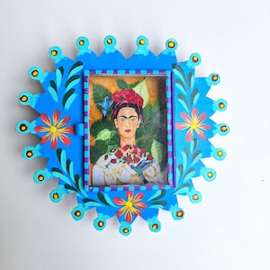 May include: A rectangular wall frame with a bright blue sunburst border featuring painted floral motifs in red, yellow, and green. The center contains a portrait of a person with dark hair, a red floral headpiece, and a white garment with red flowers.