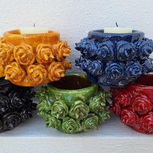 May include: Five ceramic candle holders with a rose design. The holders are in different colors: yellow, blue, green, red, and black.