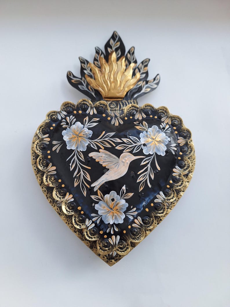 Puede incluir: Adorno de pared en forma de coraz&oacute;n pintado a mano. La base negra est&aacute; adornada con dise&ntilde;os florales blancos y dorados y un colibr&iacute;. Los bordes est&aacute;n adornados con filigrana dorada. Un dise&ntilde;o de llama dorada y negra remata el coraz&oacute;n.